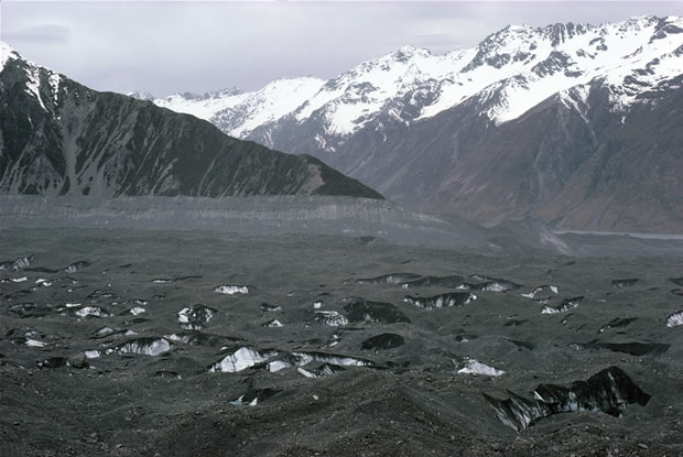 Tasman Glacier: surface and lateral moraine in eastward crossview to Malte-Brun Range (left) and Liebig Range (right) with Murchison Glacier entering between. The ice exposed here extends vertically hundreds of meters to the glacier valley floor. The movement of the ice mass is from left to right.