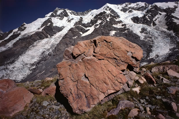 Weathered rocks on De La Beche ridge which separates the Tasman and Rudolph glaciers. Two steep glaciers on the Main Divide in background. The red colouration of the surface of the rocks shown here takes 300-400 years of iron-oxidative weathering to develop.