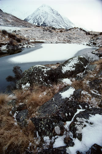 Tasman Valley: southward view of west-side tarn in winter with lichen-covered rock and sub-alpine vegetation including tussock and the dark, tangled and prickly New Zealand native shrub, Matagouri.