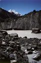 Tasman Glacier terminal face blackened by surface moraine rockfall, melting in summer sun. Glacier rock-flour whitens the terminal lake and the moraine boulders in the foreground. Mt Cook in far distance.